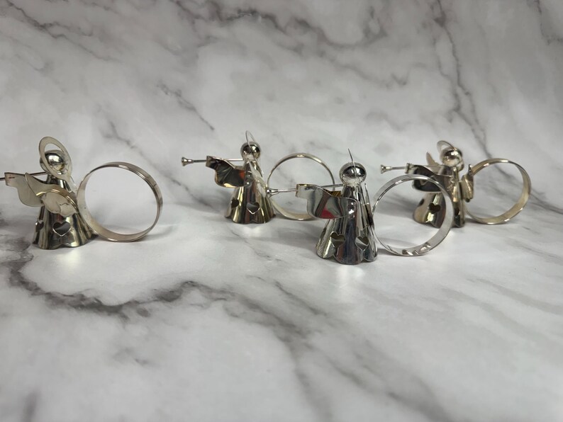 May include: Silver angel napkin rings. Each angel figure is playing a trumpet and has a halo. The napkin rings are made of metal and have a circular design. The angels are standing on a white and gray marble surface.