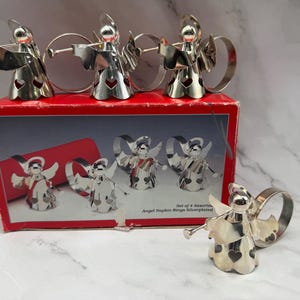 May include: A set of four silver-plated angel napkin rings, each with a trumpet and heart-shaped cutouts. The napkin rings are displayed on a red box with an illustration of the angel napkin rings. The box reads "Angel Napkin Rings Silverplated."