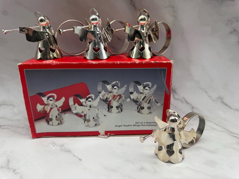 May include: A set of four silver-plated angel napkin rings, each with a trumpet and heart-shaped cutouts. The napkin rings are displayed on a red box with an illustration of the angel napkin rings. The box reads "Angel Napkin Rings Silverplated."