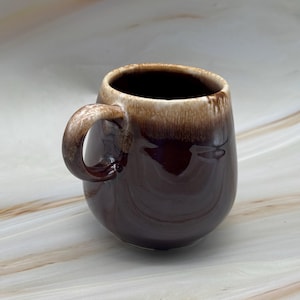 Puede incluir: Una taza de cerámica marrón con un cuerpo redondeado y un asa curvada. La taza tiene un exterior marrón oscuro que se desvanece a un marrón más claro en el borde. El interior es marrón oscuro. La taza está diseñada para bebidas calientes.