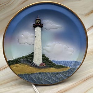 Może przedstawiać: Okrągła dekoracja ścienna przedstawiająca białą latarnię morską na wyspie, na tle błękitnego nieba i wody. Latarnia ma czerwony szczyt i jest otoczona zielenią. Ma złotą obwódkę i biały przewód elektryczny. Średnica około 20 cm.