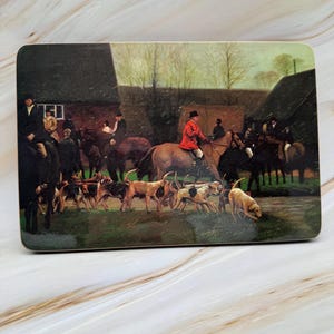 May include: Rectangular coaster with a vintage hunting scene. The image shows riders on horseback, hounds, and a rural setting with buildings and trees. The riders are wearing traditional attire, with one in a red coat.