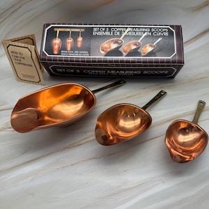 May include: A set of three copper measuring scoops with brass handles, displayed with their original box and a care guide. The box reads "Set of 3 Copper Measuring Scoops". The scoops are different sizes and have a shiny finish.