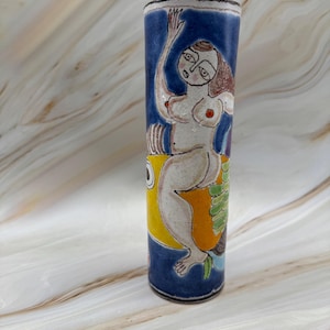 May include: A cylindrical ceramic vase featuring a hand-painted illustration of a figure with brown hair sitting atop a large yellow fish. The background is a deep blue, and the figure is depicted in a light cream tone with red accents.