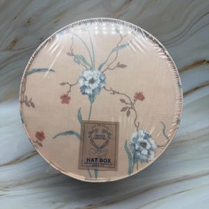 May include: Round hat box with a floral design in peach, blue, and red. The box is wrapped in clear plastic. A label reads "French Country Hat Box."