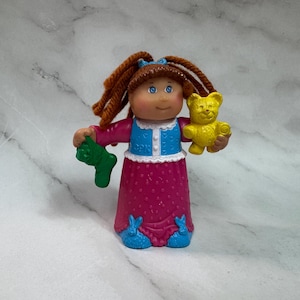 Può includere: Una statuina vintage della bambola Cabbage Patch Kids. La bambola ha capelli di filo marrone, occhi blu e indossa un vestito rosa a pois bianchi, un gilet blu e scarpe blu. Tiene in mano una calza verde e un orsacchiotto giallo.