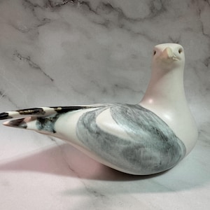 Op de afbeelding: Een keramische zeemeeuwsculptuur in rusthouding. De vogel is voornamelijk wit met grijze en zwarte markeringen op de vleugels en staart. De sculptuur heeft een gladde, glanzende afwerking, op een marmeren achtergrond.