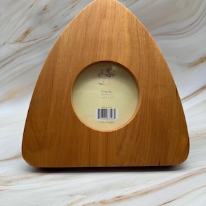May include: A wooden picture frame with a unique triangular shape and a circular opening. The frame is made of light-colored wood and has a natural finish. The opening is sized at 3.5 x 3.5 inches. The frame has a minimalist design.