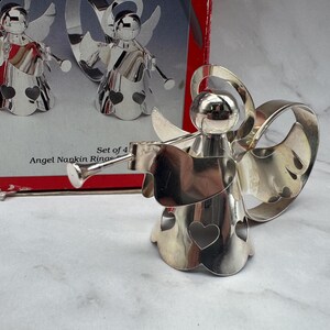 May include: Set of four silver-colored angel napkin rings. Each angel figure has wings, a heart-shaped cutout, and a trumpet. The box reads "Angel Napkin Rings". The angels are displayed on a white and gray surface.