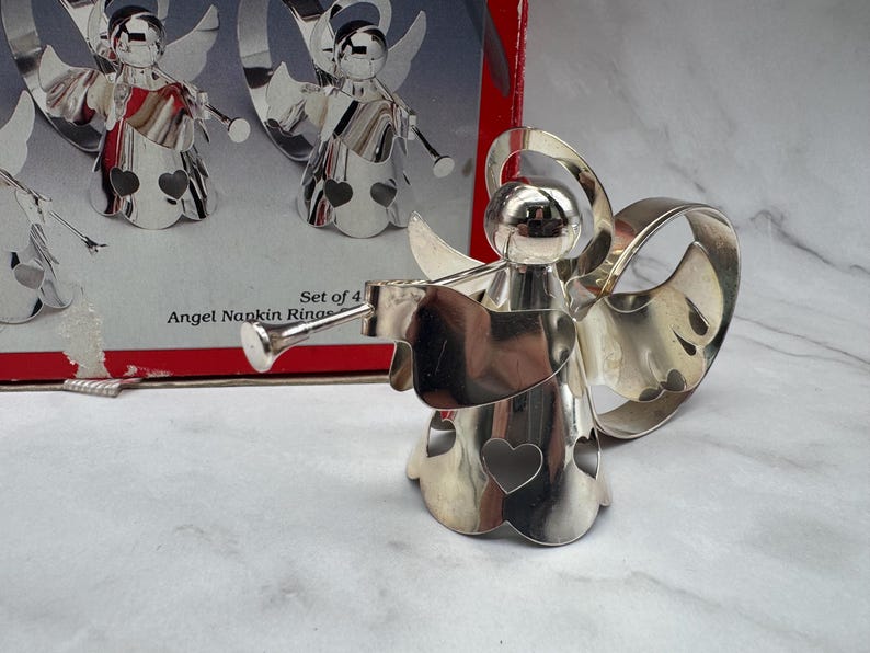 May include: Set of four silver-colored angel napkin rings. Each angel figure has wings, a heart-shaped cutout, and a trumpet. The box reads "Angel Napkin Rings". The angels are displayed on a white and gray surface.