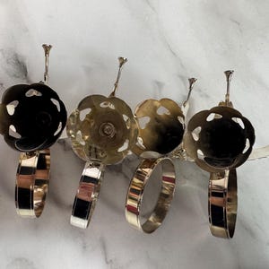 May include: Four silver-toned napkin rings with a floral design. Each ring has a circular band and a decorative top with a flower-like shape. The flower tops have cut-out heart shapes. The napkin rings are displayed on a white marble surface.