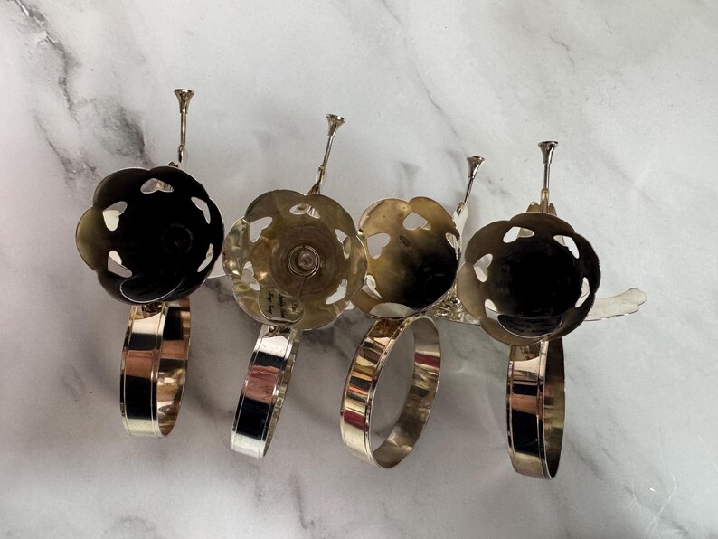 May include: Four silver-toned napkin rings with a floral design. Each ring has a circular band and a decorative top with a flower-like shape. The flower tops have cut-out heart shapes. The napkin rings are displayed on a white marble surface.