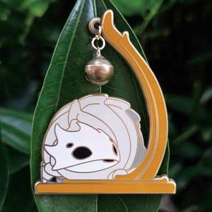 Könnte beinhalten: Ein weiß-grauer Emaille-Pin mit einem stilisierten Kreaturen-Design, eingebettet in einen goldfarbenen Rahmen. Eine kleine goldene Glocke hängt von oben. Der Pin wird auf einem grünen Blatt vor einem verschwommenen grünen Hintergrund präsentiert.