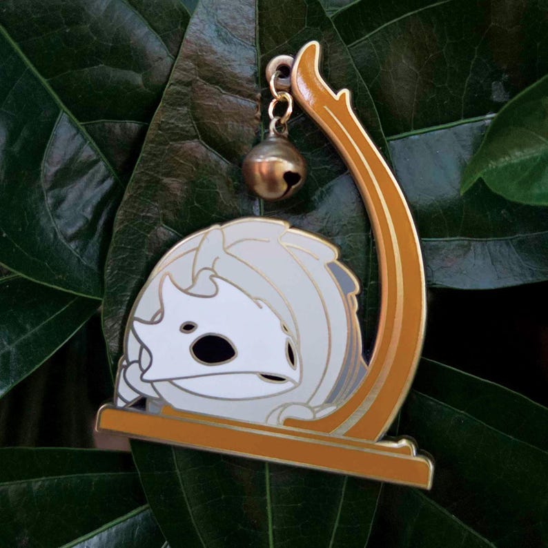 May include: A white and gray enamel pin featuring a stylized creature with a bell charm. The pin has a gold-colored frame and a small bell hanging from a chain. The pin is set against a dark green leaf.