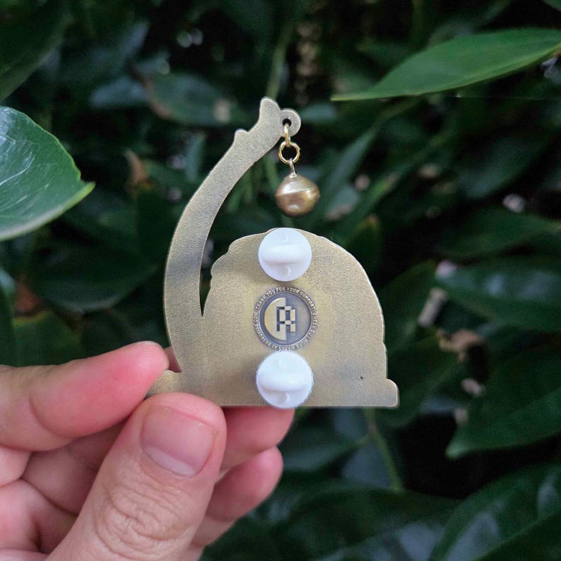 May include: A bronze-colored metal pin with a curved design, a small gold-colored ball, and two white pin backs. The pin features a circular emblem with a logo. The pin is held against a backdrop of green leaves.