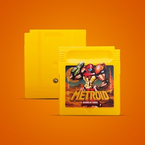 Puede incluir: Un cartucho de videojuego amarillo Metroid Return of Samus con una colorida ilustración de un personaje con un traje rojo y amarillo. El cartucho está sobre un fondo naranja, con un segundo cartucho ligeramente detrás.