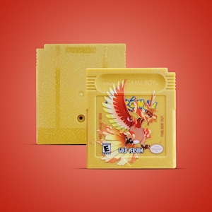 以下が含まれることがあります： 2つの黄色のゲームボーイカートリッジ。前面のカートリッジには、「Pokémon Gold Version」のテキストとともに、フェニックスのような生き物のカラフルなイラストが描かれています。もう一方のカートリッジは無地の黄色です。