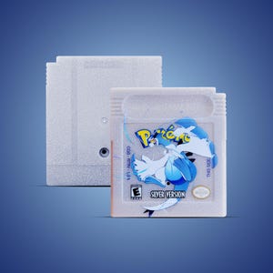 May include: A light gray Game Boy cartridge labeled "Silver Version" with a blue and white graphic. The cartridge is next to a matching gray cartridge.