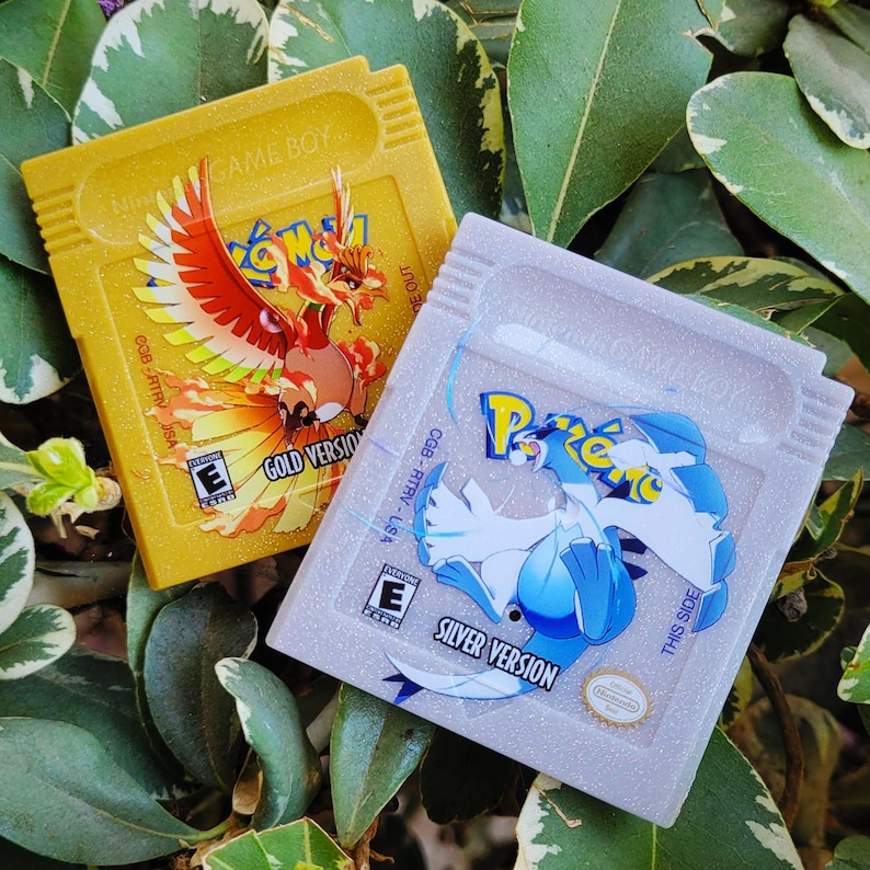 May include: Two Game Boy cartridges: a gold cartridge with a fiery bird graphic labeled "Gold Version" and a silver cartridge with a blue and white creature graphic labeled "Silver Version". Both cartridges have the "E" rating.