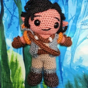 May include: A handmade crochet doll with black hair, large black eyes, and a smiling expression. The doll wears a gray vest, brown pants, and a brown strap across the chest. The doll's arms and legs are brown and orange.