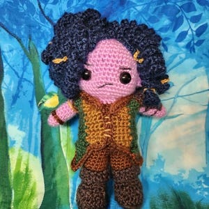 May include: A handmade crochet doll with a purple face, dark blue curly hair, and a stern expression. The doll wears a brown and green vest, brown pants, and boots. The background is a blue fabric with a tree and bird design.
