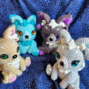 May include: Four crocheted stuffed animals in various colors, including light blue, gray, and brown. The animals have large eyes and are sitting on a blue fabric background.