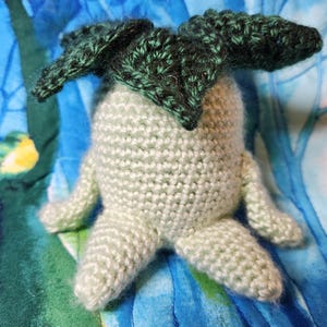 May include: A handmade crochet doll in the shape of a mandrake plant. The doll has a light green body with small arms and legs. The top of the doll features dark green leaves. The doll is set against a blue and green patterned background.