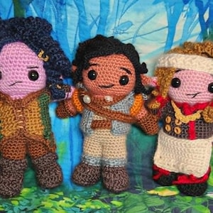 May include: Three handmade crochet dolls with detailed features. The dolls have pink skin, black eyes, and various outfits in brown, blue, and beige. The background is a watercolour painting with blue and green hues.