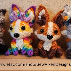 May include: Four handmade crochet fox plushies in various colors. One fox has rainbow ears and legs, another has orange and white fur, and two are gray and white. The plushies have large black eyes and are sitting side by side. Text at the bottom reads: www.etsy.com/shop/SewVixenDesigns.