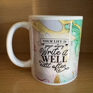 Puede incluir: Taza de cerámica blanca con asa curva. La taza presenta un diseño colorido con tonos dorados, verdes y rosas. El texto en la taza dice "YOUR LIFE IS your story Write it WELL edit often."