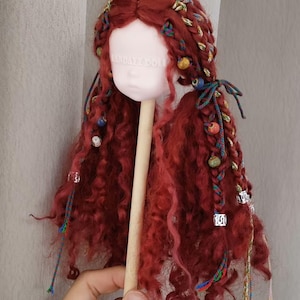 Puede incluir: Una cabeza de muñeca con pelo largo, rizado y rojo peinado con trenzas y cuentas de colores. La cabeza de la muñeca está montada en un palo de madera. La cabeza de la muñeca tiene una tez pálida.