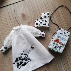 May include: A white doll dress with a black and white cow design, matching cow-print hat, and a small purse with a cow and farm scene. The dress has long sleeves with cow-print cuffs.