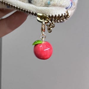 May include: A red apple charm with a green leaf, suspended from a gold-coloured zip. The zip is attached to a cream-coloured, textured fabric with flecks of colour. The charm is a decorative accessory.