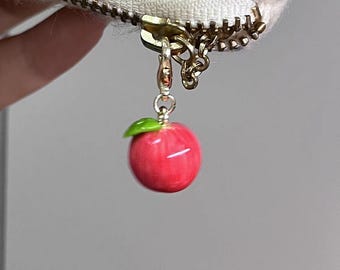 May include: A red apple charm with a green leaf, suspended from a gold-coloured zip. The zip is attached to a cream-coloured, textured fabric with flecks of colour. The charm is a decorative accessory.