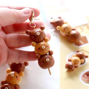 Könnte beinhalten: Ein handgefertigter Schlüsselanhänger mit drei bärenförmigen Ornamenten auf einem Holzspieß. Die Bären sind in verschiedenen Braun- und Beigetönen gehalten und haben kleine weiße Akzente für Augen und Nasen. Ein goldfarbener Verschluss ist am obersten Bären befestigt.