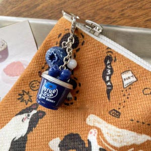 Può includere: Un ciondolo a forma di tazza blu con la scritta "Blue Cup Blue Berry" è attaccato a una catena d'argento. Il ciondolo è decorato con mirtilli e perline bianche. Il ciondolo è attaccato a una chiusura argentata.