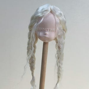 Puede incluir: Una cabeza de muñeca con cabello largo, ondulado y rubio claro peinado en dos trenzas. La cara de la muñeca es pálida con el texto "LANDAZZ DOLL" impreso en la frente. La cabeza está unida a una clavija de madera.