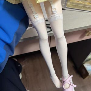 Op de afbeelding: De benen van een pop zijn te zien, met witte kousen die worden vastgehouden door bretels. De pop heeft roze Mary Jane schoenen met een witte zool. De pop draagt een gele en roze rok.