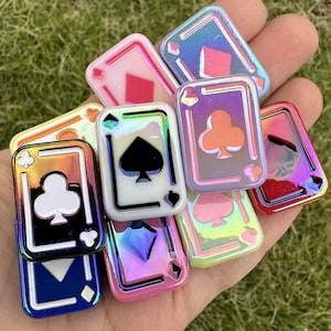 May include: A collection of iridescent playing card charms in various colors, including red, blue, and green. Each charm features a different suit symbol: hearts, diamonds, clubs, and spades. The charms have a glossy finish and are held in a hand.