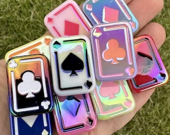 May include: A collection of iridescent playing card charms in various colors, including red, blue, and green. Each charm features a different suit symbol: hearts, diamonds, clubs, and spades. The charms have a glossy finish and are held in a hand.