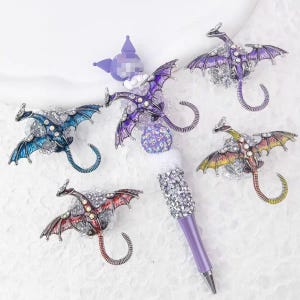 Puede incluir: Un bolígrafo decorativo con un cuerpo morado y un diseño de dragón. El bolígrafo presenta múltiples adornos de dragones en varios colores, incluyendo azul, morado, rojo y amarillo, cada uno adornado con pedrería. La parte superior del bolígrafo está adornada con una bola de cristal y una corona morada.