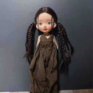 Puede incluir: Una muñeca artesanal con largas trenzas castañas oscuras y una expresión serena. La muñeca viste un vestido marrón fluido y zapatos a juego. La muñeca es una creación artística única.