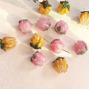 Puede incluir: Una colección de cuentas de flores de vidrio en tonos amarillos y rosas. Las cuentas están detalladas con acentos verdes, que se asemejan a diseños florales en miniatura. Las cuentas están dispersas sobre una superficie clara.