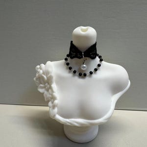 May include: A white bust sculpture adorned with a black lace choker and a beaded necklace featuring a pearl pendant. The choker has a bow design. The bust has floral detailing on the shoulder.