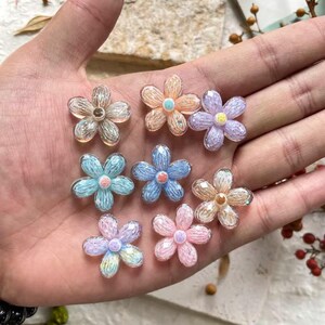 以下が含まれることがあります： 様々なパステルカラーの、半透明な花の形をした装飾品のコレクション。 ピンク、青、オレンジ、紫など、中心にビーズが付いています。 クラフトやDIYプロジェクトに最適な装飾要素です。