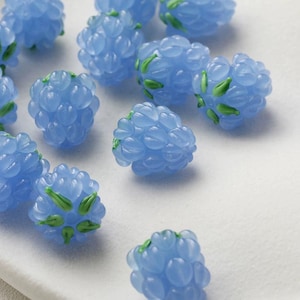 Puede incluir: Primer plano de varias cuentas en forma de bayas de color azul claro con detalles verdes. Las cuentas tienen un diseño agrupado, que se asemeja a un racimo de uvas o bayas. Las cuentas están dispersas sobre una superficie blanca.