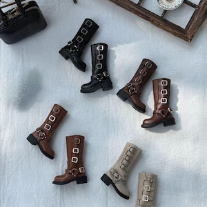 May include: A collection of miniature boots in various colors, including black, brown, and beige. Each boot features multiple buckles and straps, with a chunky heel. The boots are arranged on a light-colored surface, suggesting a fashion or craft theme.