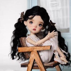 Könnte beinhalten: Porzellanpuppe mit schwarzen Locken in Zöpfen, die auf einem kleinen Holzstuhl sitzt. Die Puppe trägt ein weißes Kleid und hat blaue Augen und rosige Wangen. Braune Schleifen schmücken das Haar der Puppe.