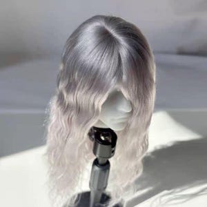 Puede incluir: Una cabeza de muñeca con una peluca plateada y blanca. La peluca tiene un cabello largo y ondulado que se desvanece de plateado a blanco en los extremos. La cabeza de la muñeca está montada en un soporte negro, sobre un fondo suave y neutro.