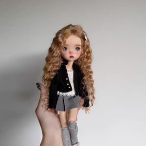 Puede incluir: Una muñeca con largo cabello castaño rojizo ondulado, ojos azules y mejillas rosadas. La muñeca viste una chaqueta negra, una blusa blanca, una falda plisada gris, calcetines altos grises y zapatos beige. Un pasador blanco adorna el cabello de la muñeca.